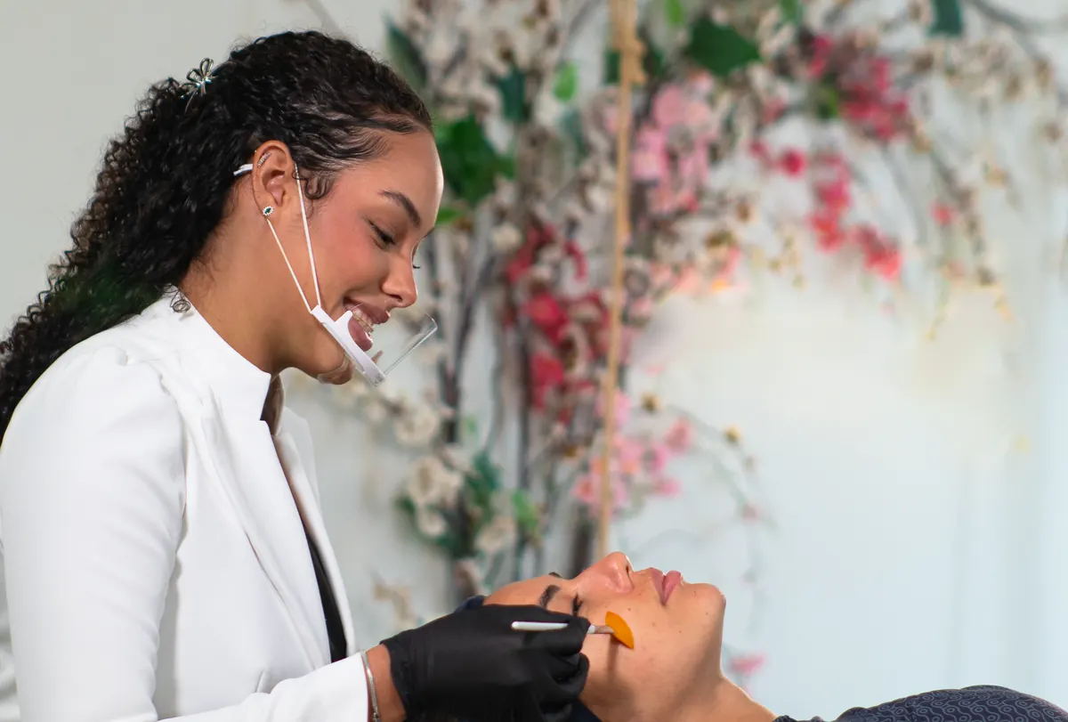 Sarah, sorrindo, aplica um peeling com pincel no rosto de uma cliente